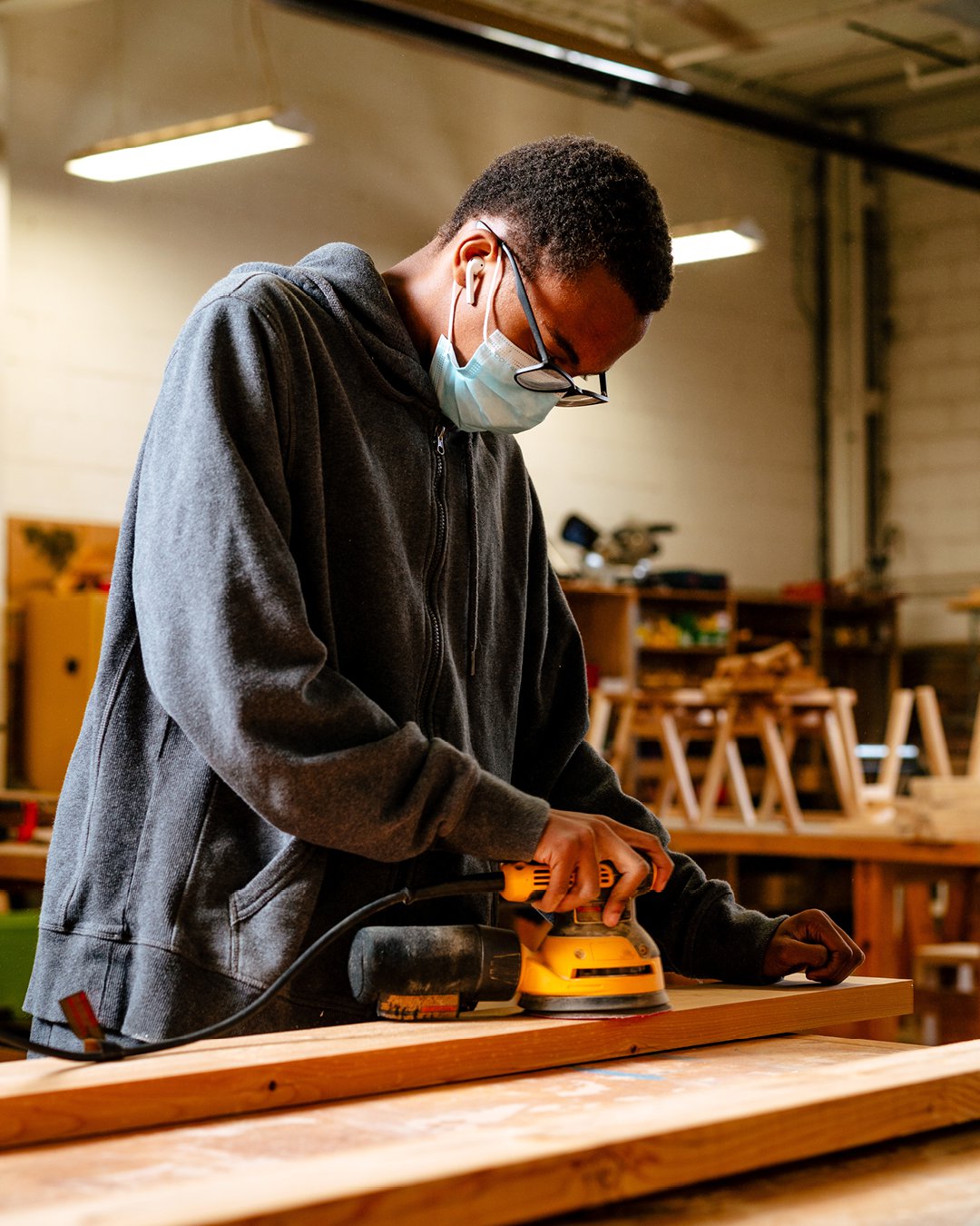 A student at the Brightmoor Maker Space standing at a work table sanding a wooden plank in the wood shop, Kazam! Magazine.
